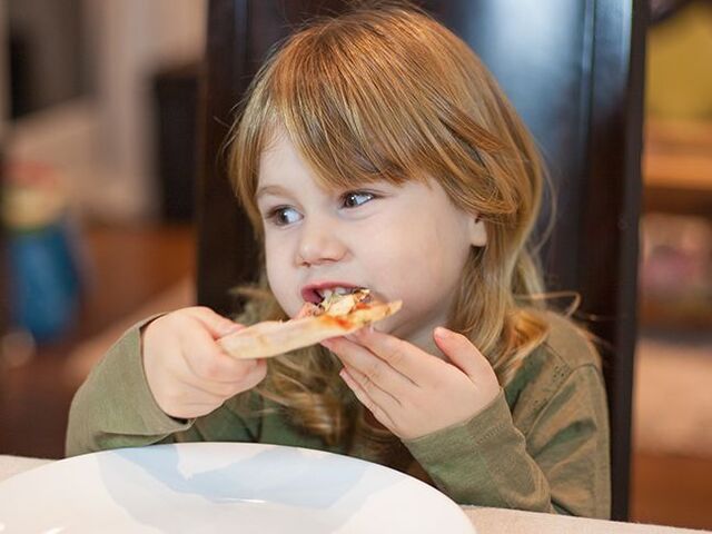 Una bimba a tavola morde un trancio di pizza.