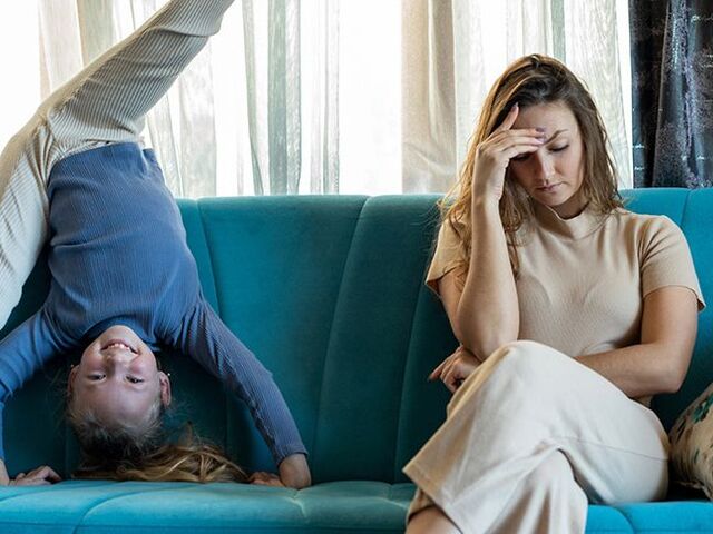 Una mamma siede irritata sul divano, mentre la figlia fa ginnastica accanto a lei.