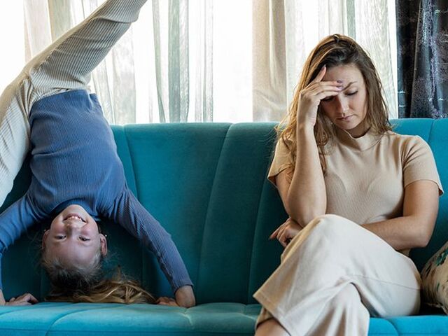 Una mamma siede irritata sul divano, mentre la figlia fa ginnastica accanto a lei.